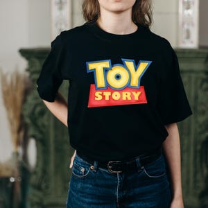 May include: A black short-sleeved t-shirt featuring the Toy Story logo printed on the chest. The logo displays the word TOY in large yellow letters with blue outlines above the word STORY in yellow letters on a red rectangular background.