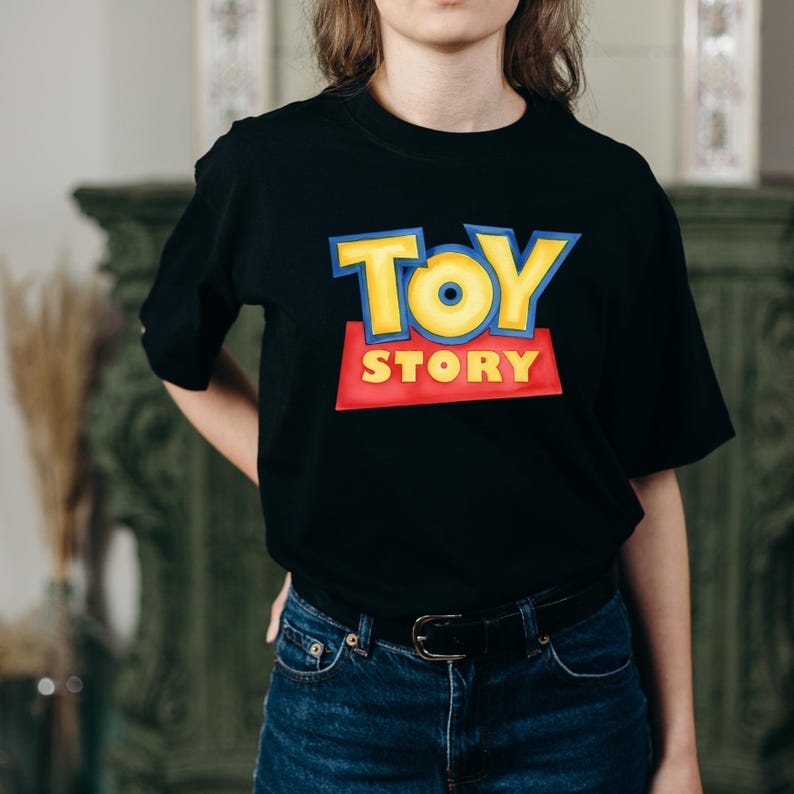 May include: A black short-sleeved t-shirt featuring the Toy Story logo printed on the chest. The logo displays the word TOY in large yellow letters with blue outlines above the word STORY in yellow letters on a red rectangular background.