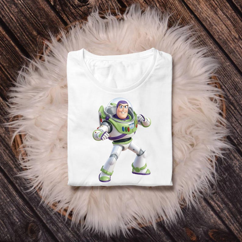May include: A white short-sleeved t-shirt featuring a centered graphic of the character Buzz Lightyear from Toy Story. The character is depicted in a standing pose wearing a white, green, and purple space suit with a clear helmet dome.