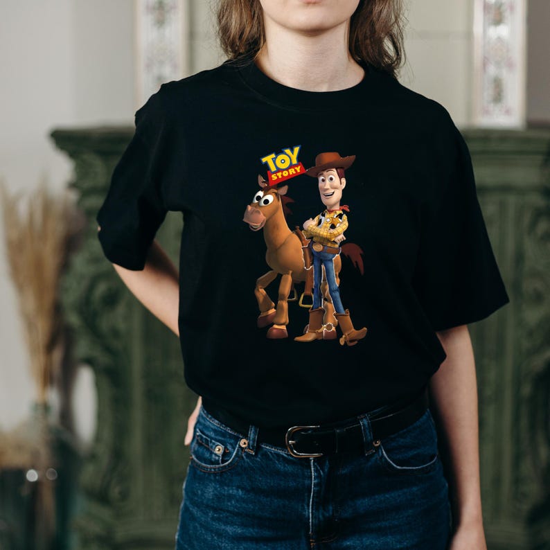 May include: A black short-sleeved t-shirt featuring a graphic print of the characters Woody and Bullseye from Toy Story. The design includes the colourful Toy Story logo positioned above the characters, who are standing together in their classic attire.