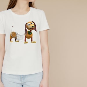 May include: A white short-sleeved t-shirt featuring a graphic print of a brown and tan dachshund toy with a metal spring body, long ears, and a green collar. The dog character has a smiling expression with large eyes and a black nose.