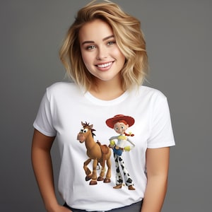 May include: A white short-sleeved t-shirt featuring a graphic print of a brown toy horse and a female toy character wearing a red cowboy hat, a yellow shirt, blue pants, and white pants with black cow-print spots standing side by side.