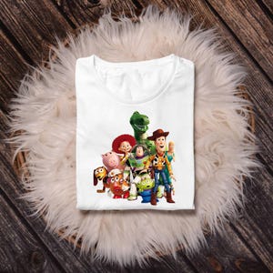 May include: A white t-shirt featuring a colourful graphic print of a group of animated characters including a cowboy, a space ranger, a green dinosaur, a pig, a potato-shaped figure, a cowgirl, a small green alien, and a blonde doll.