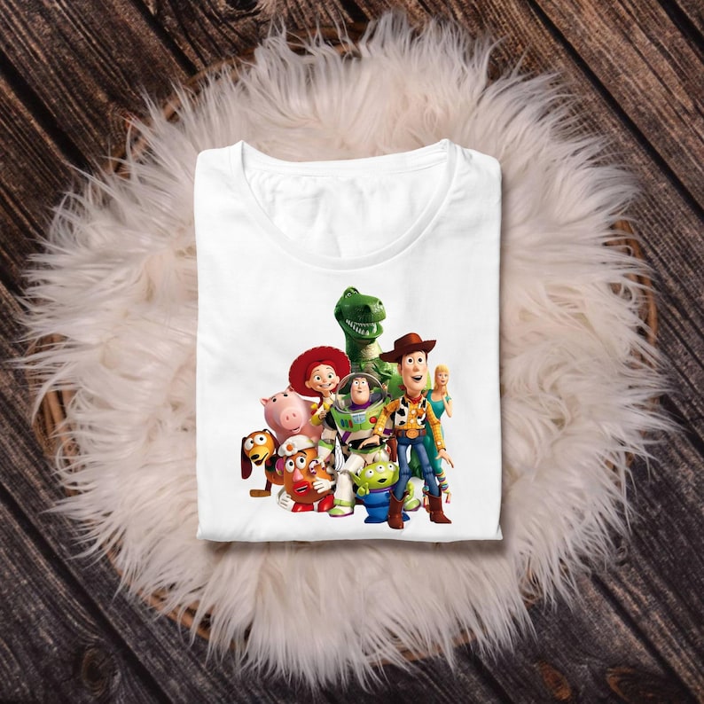 May include: A white t-shirt featuring a colourful graphic print of a group of animated characters including a cowboy, a space ranger, a green dinosaur, a pig, a potato-shaped figure, a cowgirl, a small green alien, and a blonde doll.