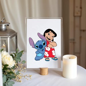 May include: A rectangular clear acrylic frame standing on a small wooden base, displaying a printed illustration of the blue alien character Stitch standing next to a girl with black hair wearing a red dress with a white leaf pattern.