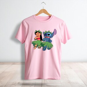 May include: A light pink short-sleeved t-shirt featuring a graphic print of Lilo and Stitch wearing green leaf skirts and headpieces. The characters are depicted in a hula dancing pose. The shirt has a crew neckline and is displayed on a hanger.