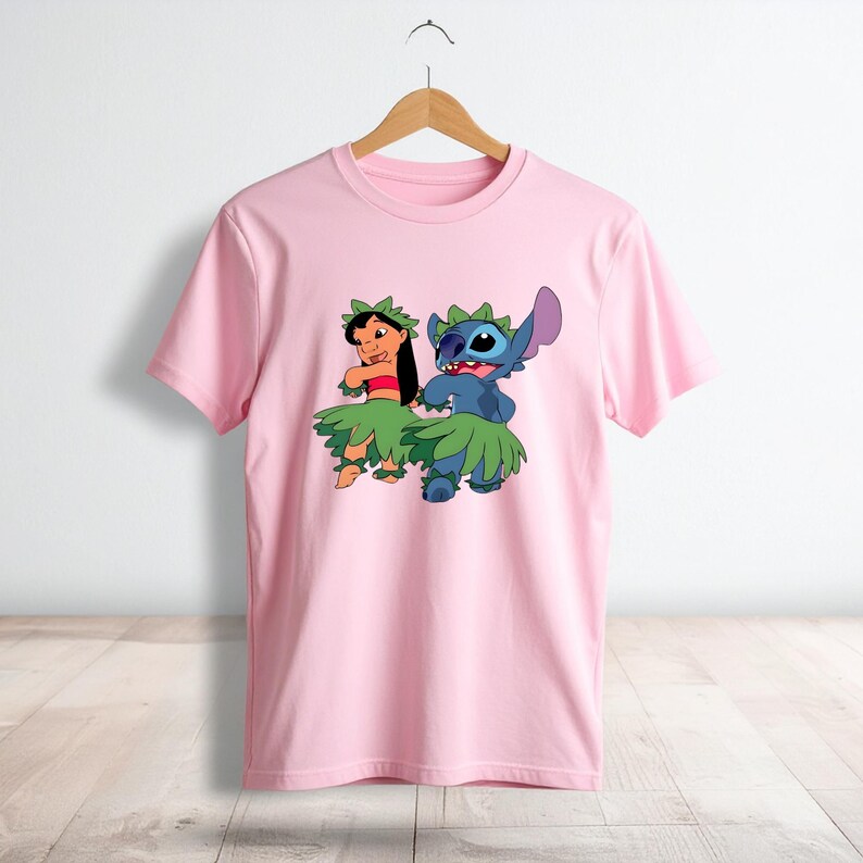 May include: A light pink short-sleeved t-shirt featuring a graphic print of Lilo and Stitch wearing green leaf skirts and headpieces. The characters are depicted in a hula dancing pose. The shirt has a crew neckline and is displayed on a hanger.
