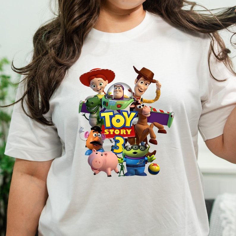 May include: A white short-sleeved t-shirt featuring a colourful graphic print of the Toy Story 3 movie characters, including Woody, Buzz Lightyear, Jessie, Rex, Mr. Potato Head, Hamm, Bullseye, and an Alien, with the Toy Story 3 logo centred.