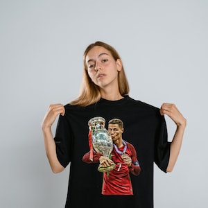 May include: A black short-sleeved t-shirt featuring a graphic print of a soccer player in a red jersey holding a large silver trophy. The player is wearing a blue medal around their neck and is shown kissing the trophy. The design is centered on the chest.
