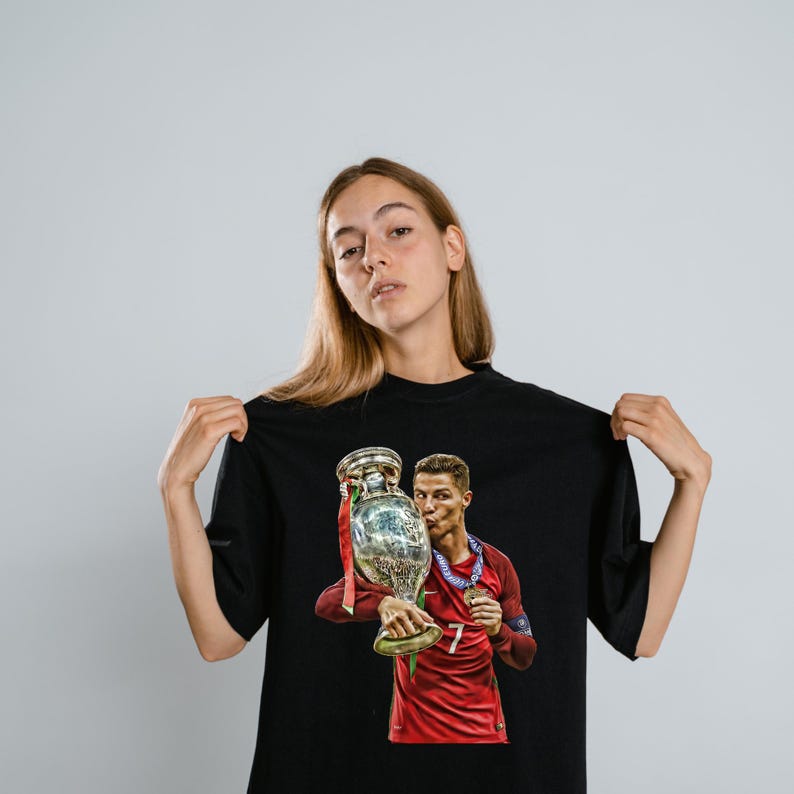 May include: A black short-sleeved t-shirt featuring a graphic print of a soccer player in a red jersey holding a large silver trophy. The player is wearing a blue medal around their neck and is shown kissing the trophy. The design is centered on the chest.