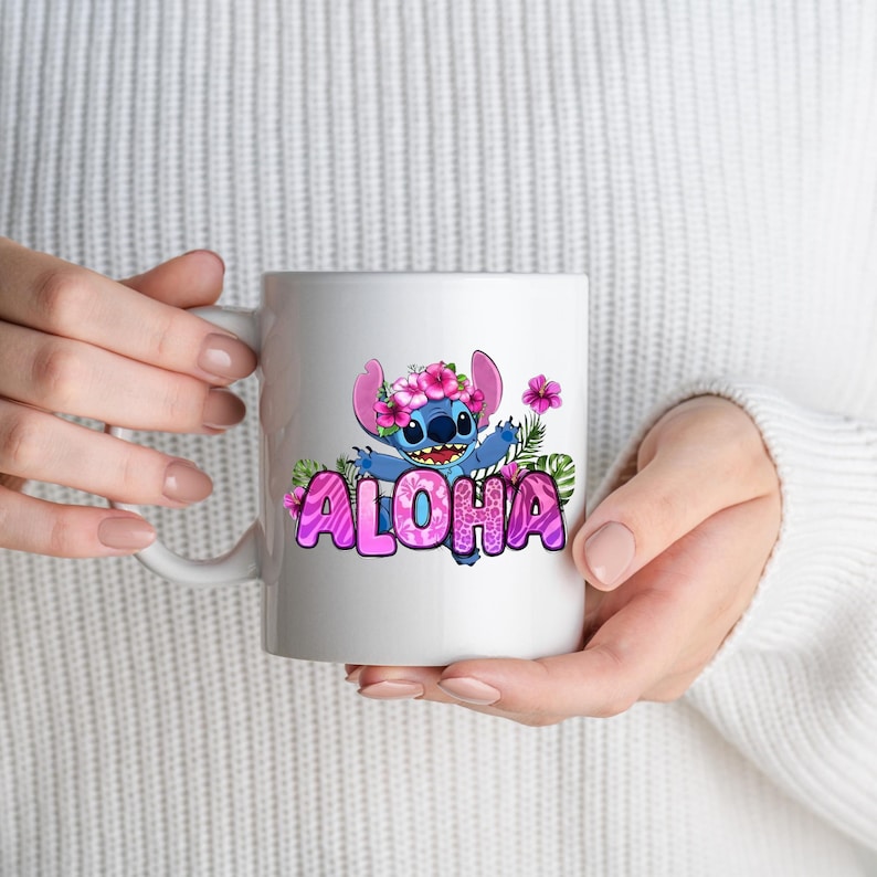 May include: A white ceramic mug featuring a graphic of a blue alien character wearing a pink hibiscus flower crown. The character is surrounded by green tropical leaves and pink flowers, with the word ALOHA in large, pink, patterned letters.
