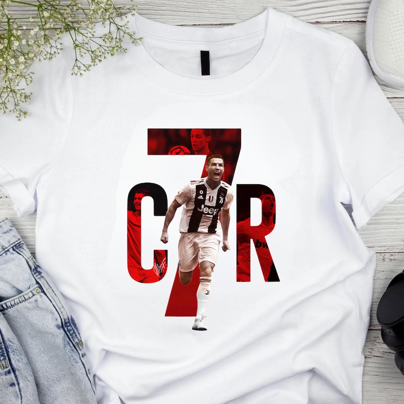 May include: A white short-sleeved t-shirt featuring a large graphic print of the number 7 and the letters CR. The design incorporates a photographic image of a soccer player in a black and white jersey, along with red-toned background elements.