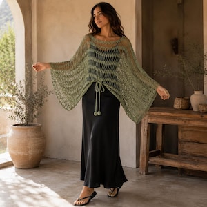 May include: An olive green crochet poncho with a wide, open-knit pattern and a drawstring waist featuring two round tassels. The poncho is worn over a black sleeveless slip dress that reaches the ankles, paired with simple black flip-flop sandals.