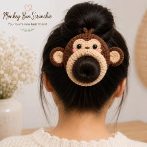 May include: A brown and beige crocheted monkey hair scrunchie featuring a circular face design with small ears, black eyes, and pink cheeks. The text Monkey Bun Scrunchie and Your bun's new best friend is printed in the upper left corner of the image.