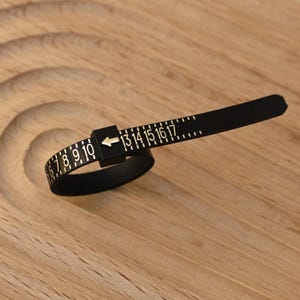 May include: A black plastic ring sizer tool featuring a belt-like design with white printed numbers ranging from 6 to 17. The tool includes a square buckle component with a white arrow pointing toward the numerical scale for measuring finger size.