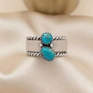 May include: A silver ring featuring two oval turquoise stones set vertically. The band consists of a smooth central silver strip flanked by two twisted rope-style silver borders. The stones are a vibrant blue-green with dark matrix inclusions.