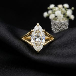 May include: A gold ring featuring a large, marquise-cut clear gemstone held by a six-prong setting. The band has a split-shank design that tapers toward the center stone, creating a sleek and structured appearance with a polished metallic finish.