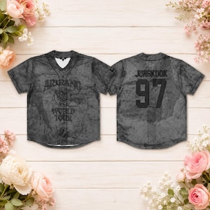 Puede incluir: Una camiseta de béisbol de manga corta con botones y un patrón de mármol gris oscuro. El frente presenta el texto "AROUND THE WORLD TOUR" con un gráfico circular. La parte posterior muestra el nombre "JUNGKOOK" sobre el gran número negro "97".