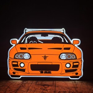 Könnte beinhalten: Ein orangefarbenes LED-Neonschild in Form der Vorderansicht eines Sportwagens. Das Auto verfügt über einen Heckspoiler, runde Scheinwerfer, ein zentrales Emblem und die auf die vordere Stoßstange gedruckte Nummer 12. Das Schild leuchtet hell vor einem dunklen Hintergrund.
