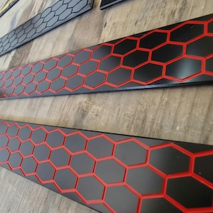 Può includere: Tre lunghe strisce rettangolari caratterizzate da una superficie nera con un motivo a nido d'ape rosso incassato. Le strisce sono disposte in parallelo, mostrando un design a griglia esagonale ripetitivo che crea un netto contrasto.