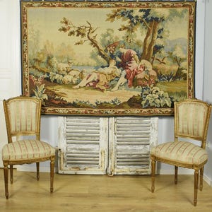 May include: A large rectangular wall tapestry featuring a pastoral scene with two figures resting under a tree near sheep, framed by a floral border. Two wooden chairs with striped fabric upholstery and gold-toned frames are positioned in front.