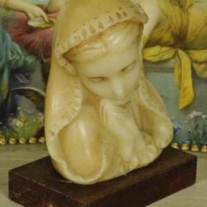 K&ouml;nnte beinhalten: Eine cremefarbene Wachsskulptur einer Figur mit Kopfbedeckung, deren H&auml;nde in einer gebets&auml;hnlichen Geste gefaltet sind. Die Skulptur ist auf einem rechteckigen, dunkelbraunen Holzsockel montiert und weist eine glatte, geformte Oberfl&auml;che auf.