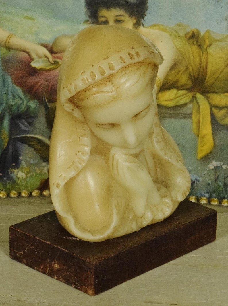 K&ouml;nnte beinhalten: Eine cremefarbene Wachsskulptur einer Figur mit Kopfbedeckung, deren H&auml;nde in einer gebets&auml;hnlichen Geste gefaltet sind. Die Skulptur ist auf einem rechteckigen, dunkelbraunen Holzsockel montiert und weist eine glatte, geformte Oberfl&auml;che auf.