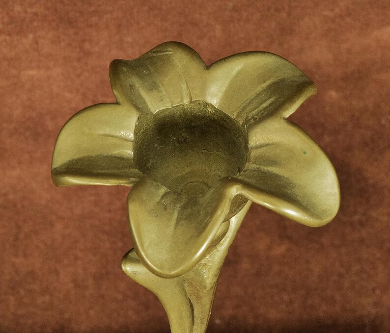 K&ouml;nnte beinhalten: Ein Kerzenhalter aus Messing in Form einer f&uuml;nfbl&auml;ttrigen Blume. Das Metall hat eine glatte, matte Oberfl&auml;che mit einem goldenen Farbton. Die Mitte der Blume weist eine kreisf&ouml;rmige &Ouml;ffnung auf, die f&uuml;r eine Stabkerze ausgelegt ist.