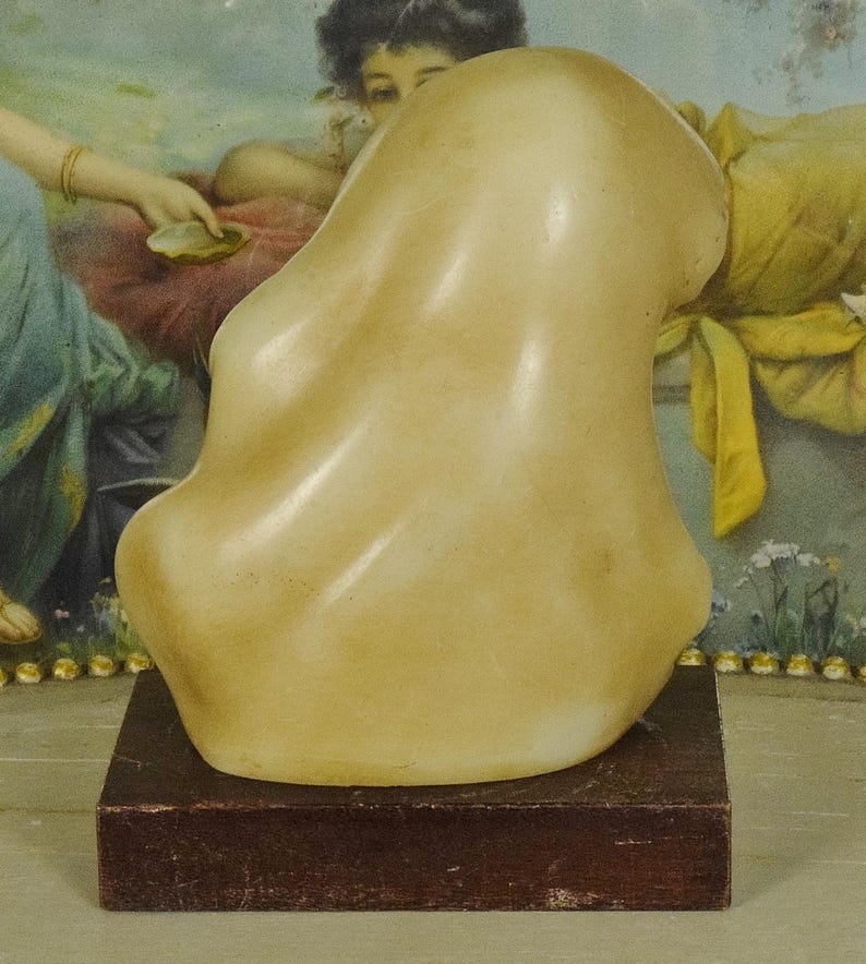 K&ouml;nnte beinhalten: Eine glatte, cremefarbene abstrakte Steinskulptur mit organischen, flie&szlig;enden Kurven. Die Skulptur ist auf einem rechteckigen, dunkelbraunen Holzsockel montiert, der der polierten, hellen Steinform eine stabile Basis verleiht.