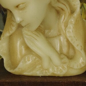 K&ouml;nnte beinhalten: Eine cremefarbene Wachskerze, geformt als B&uuml;ste einer Figur mit geschlossenen Augen und gefalteten H&auml;nden in Gebetshaltung. Die Figur tr&auml;gt eine drapierte Kopfbedeckung mit strukturiertem Rand, die auf einem dunkelbraunen rechteckigen Sockel steht.