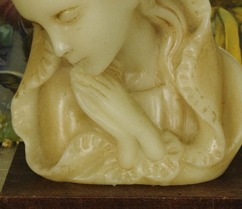 K&ouml;nnte beinhalten: Eine cremefarbene Wachskerze, geformt als B&uuml;ste einer Figur mit geschlossenen Augen und gefalteten H&auml;nden in Gebetshaltung. Die Figur tr&auml;gt eine drapierte Kopfbedeckung mit strukturiertem Rand, die auf einem dunkelbraunen rechteckigen Sockel steht.