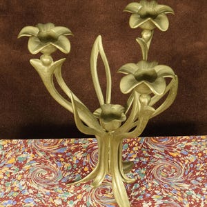 K&ouml;nnte beinhalten: Ein goldfarbener Metallkandelaber mit vier Kerzenhaltern in Form von offenen Bl&uuml;ten. Die Struktur wird von einem zentralen Stiel getragen, der sich in geschwungene, blattartige Formen verzweigt und ein botanisches Design mit glatter Oberfl&auml;che bildet.
