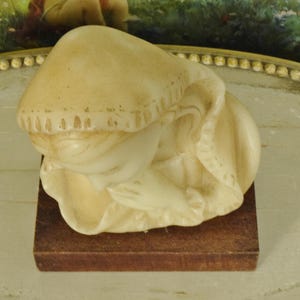 K&ouml;nnte beinhalten: Eine cremefarbene Figur einer Person mit geneigtem Kopf und in Gebetshaltung gefalteten H&auml;nden, die ein Kapuzenkleidungsst&uuml;ck mit strukturierten R&auml;ndern tr&auml;gt. Die Skulptur ist auf einem rechteckigen, dunkelbraunen Holzsockel montiert.