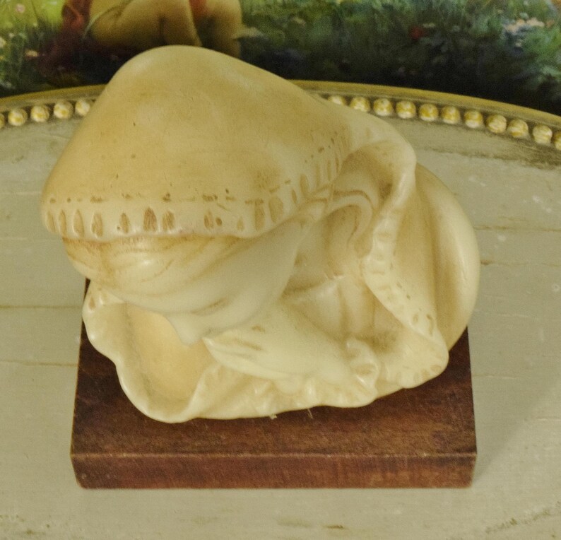 K&ouml;nnte beinhalten: Eine cremefarbene Figur einer Person mit geneigtem Kopf und in Gebetshaltung gefalteten H&auml;nden, die ein Kapuzenkleidungsst&uuml;ck mit strukturierten R&auml;ndern tr&auml;gt. Die Skulptur ist auf einem rechteckigen, dunkelbraunen Holzsockel montiert.