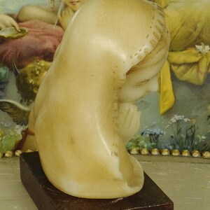 K&ouml;nnte beinhalten: Eine cremefarbene Steinb&uuml;ste einer Figur mit Kopfbedeckung, im Profil dargestellt mit gefalteten H&auml;nden in Gebetshaltung. Die Skulptur ist auf einem rechteckigen, dunkelbraunen Holzsockel montiert und weist eine glatte, polierte Oberfl&auml;che auf.