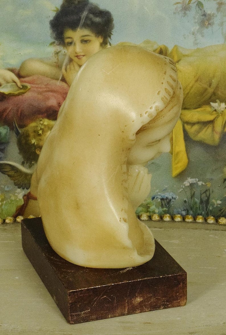 K&ouml;nnte beinhalten: Eine cremefarbene Steinb&uuml;ste einer Figur mit Kopfbedeckung, im Profil dargestellt mit gefalteten H&auml;nden in Gebetshaltung. Die Skulptur ist auf einem rechteckigen, dunkelbraunen Holzsockel montiert und weist eine glatte, polierte Oberfl&auml;che auf.