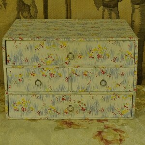 May include: A rectangular storage chest with four drawers, featuring a light-coloured surface covered in a pattern of blue grass-like blades and small red and yellow floral accents. Each drawer is equipped with a small, circular silver-toned metal pull.