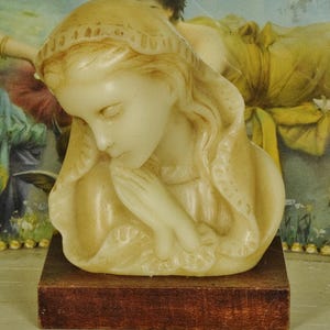 K&ouml;nnte beinhalten: Eine cremefarbene Wachsb&uuml;ste einer Figur mit geschlossenen Augen und in Gebetshaltung gefalteten H&auml;nden, die eine drapierte Kopfbedeckung mit strukturiertem Rand tr&auml;gt. Die Skulptur ist auf einem rechteckigen, dunkelbraunen Holzsockel montiert.