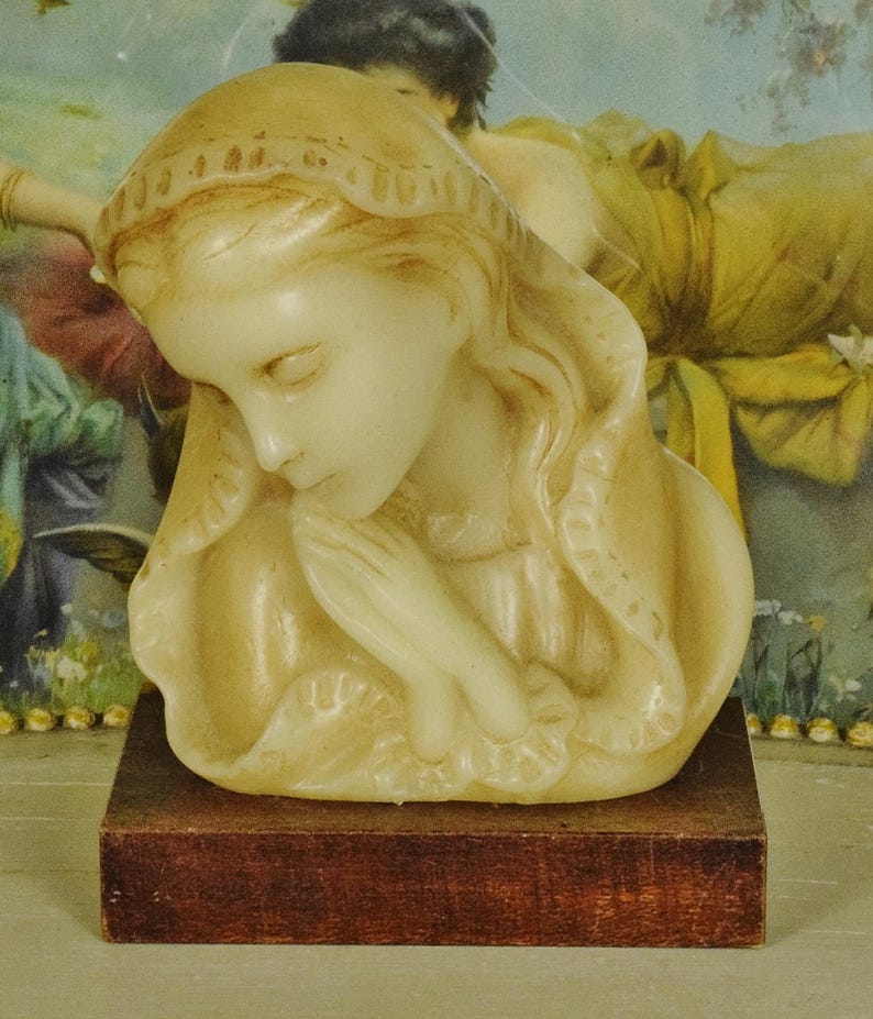 K&ouml;nnte beinhalten: Eine cremefarbene Wachsb&uuml;ste einer Figur mit geschlossenen Augen und in Gebetshaltung gefalteten H&auml;nden, die eine drapierte Kopfbedeckung mit strukturiertem Rand tr&auml;gt. Die Skulptur ist auf einem rechteckigen, dunkelbraunen Holzsockel montiert.