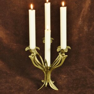 K&ouml;nnte beinhalten: Ein goldfarbener Metallkandelaber mit vier Armen in Form von stilisierten Blumenst&auml;ngeln und Bl&auml;ttern. Vier hohe, schlanke wei&szlig;e Stabkerzen stecken in den Haltern, jede mit einer kleinen, leuchtenden Flamme an der Spitze versehen.