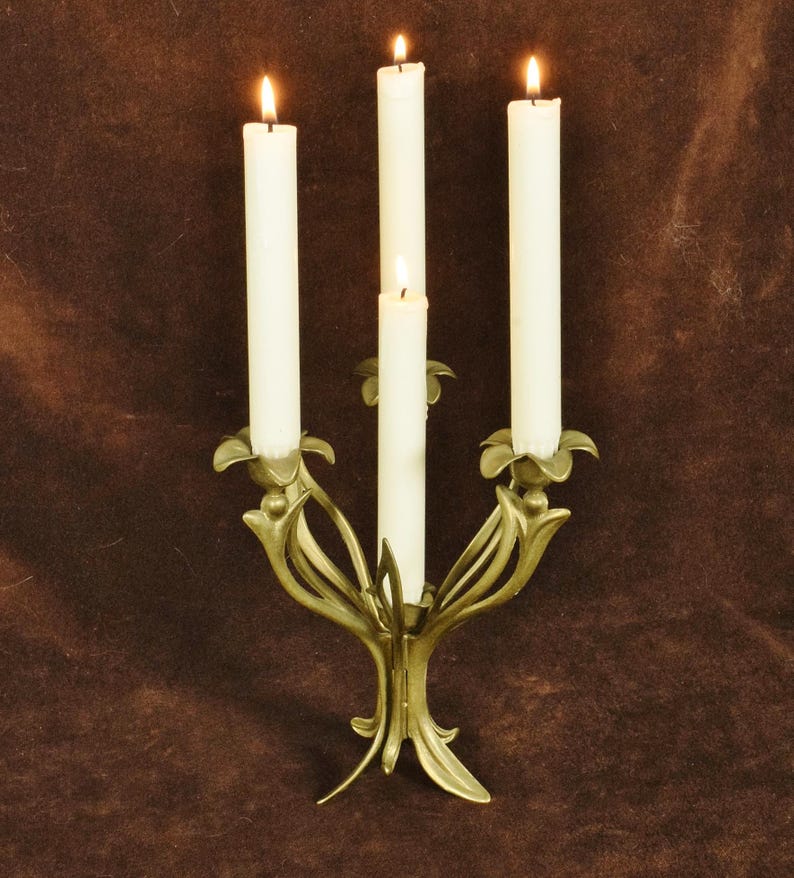K&ouml;nnte beinhalten: Ein goldfarbener Metallkandelaber mit vier Armen in Form von stilisierten Blumenst&auml;ngeln und Bl&auml;ttern. Vier hohe, schlanke wei&szlig;e Stabkerzen stecken in den Haltern, jede mit einer kleinen, leuchtenden Flamme an der Spitze versehen.