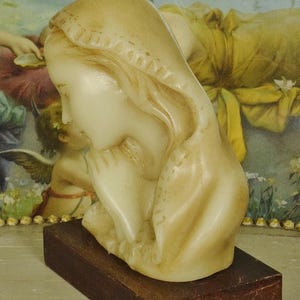 K&ouml;nnte beinhalten: Eine cremefarbene Wachsb&uuml;ste einer Figur mit Kopfbedeckung, dargestellt in einer betenden Haltung mit gefalteten H&auml;nden. Die Skulptur ist auf einem rechteckigen, dunkelbraunen Holzsockel montiert und weist glatte Konturen sowie weiche Gesichtsz&uuml;ge auf.