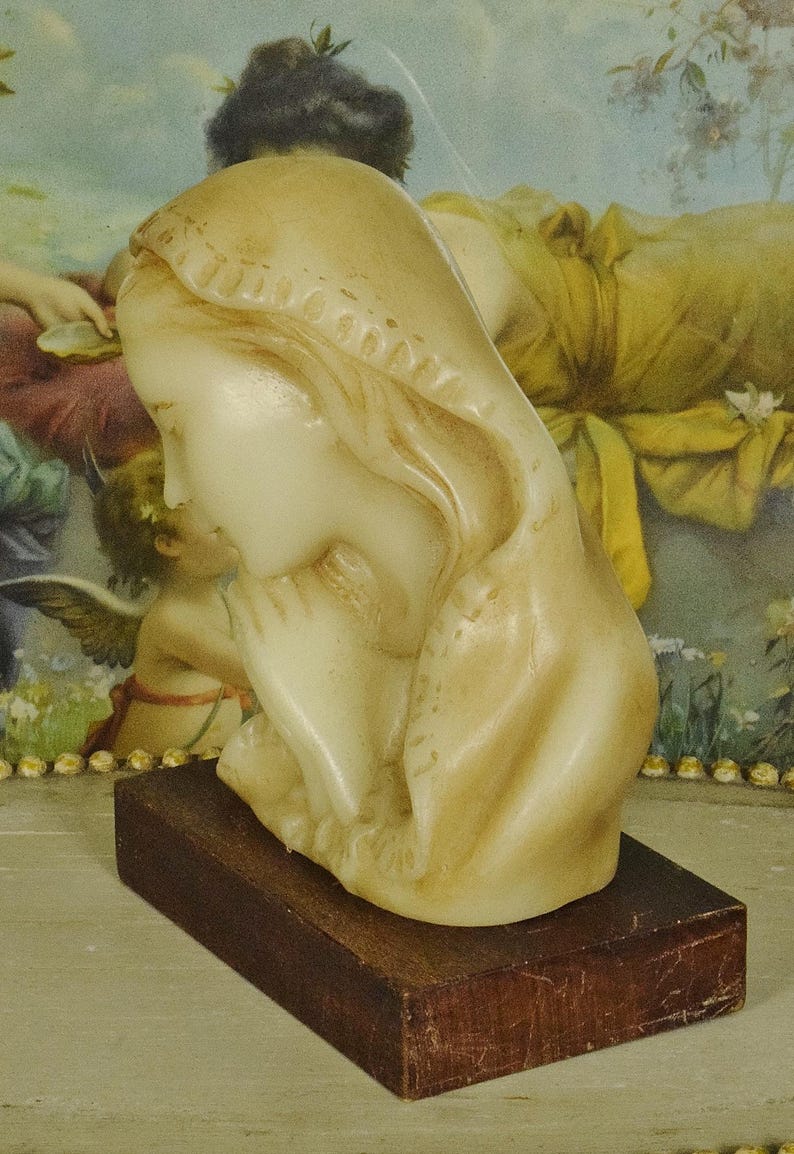 K&ouml;nnte beinhalten: Eine cremefarbene Wachsb&uuml;ste einer Figur mit Kopfbedeckung, dargestellt in einer betenden Haltung mit gefalteten H&auml;nden. Die Skulptur ist auf einem rechteckigen, dunkelbraunen Holzsockel montiert und weist glatte Konturen sowie weiche Gesichtsz&uuml;ge auf.