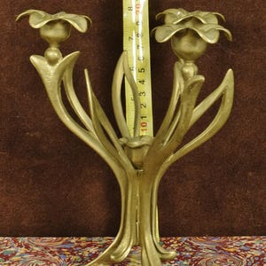 K&ouml;nnte beinhalten: Ein messingfarbener Metallkandelaber mit drei blumenf&ouml;rmigen Kerzenhaltern und geschwungenen Stielen. Ein gelbes Ma&szlig;band ist vertikal hinter der Mitte positioniert und zeigt eine H&ouml;he von etwa 24 cm f&uuml;r die Struktur an.