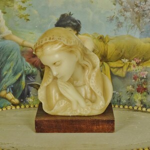 K&ouml;nnte beinhalten: Eine cremefarbene Wachsskulptur einer Person mit geneigtem Kopf und in Gebetshaltung gefalteten H&auml;nden, die eine drapierte Kopfbedeckung tr&auml;gt. Die Figur ist auf einem rechteckigen, dunkelbraunen Holzsockel montiert und zeigt glatte Details.