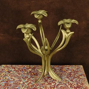 K&ouml;nnte beinhalten: Ein Messing-Kandelaber mit drei Kerzenhaltern in Form von offenen Bl&uuml;ten. Die Struktur wird von einem zentralen Stiel getragen, der sich in geschwungene, blattartige Formen verzweigt und eine botanische Silhouette mit metallischem Glanz erzeugt.