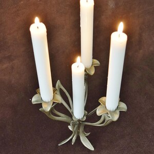 K&ouml;nnte beinhalten: Ein vierarmiger Kerzenst&auml;nder aus Metall mit mattem Goldfinish und bl&uuml;tenf&ouml;rmigen Kerzenhaltern. Vier brennende wei&szlig;e Stabkerzen stecken in den Halterungen, wobei die Flammen einen sanften Schein auf die metallische, blattartige Struktur werfen.