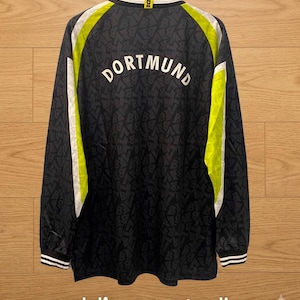 Puede incluir: Una camiseta de manga larga en negro con un patrón de letras repetidas, que presenta la palabra DORTMUND en letras blancas en la espalda. La prenda incluye paneles laterales en amarillo neón y blanco, cuello redondo y puños a rayas blancas y negras.