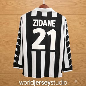 May include: A long-sleeved black and white striped sports jersey featuring the name ZIDANE and the number 21 printed in white on the back. The sleeves display a repeating white Kappa logo pattern on a black background, with a white collar.