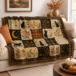 Puede incluir: Una manta tejida con diseño de patchwork que presenta gatos negros, lunas crecientes, telarañas y hojas de otoño en tonos naranja, marrón y crema. La manta tiene un borde con flecos y un patrón repetitivo de cuadros y motivos florales.